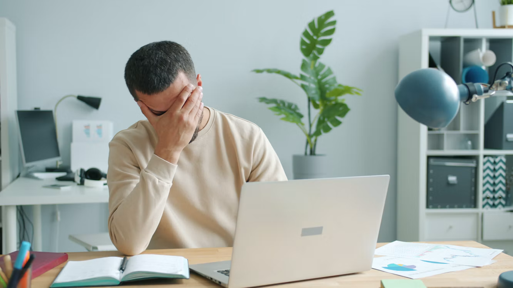 A man sat at a desk working on a laptop looking frustrated